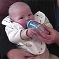A 6-8 month old baby sitting up and drinking from a bottle that is being held for them.
